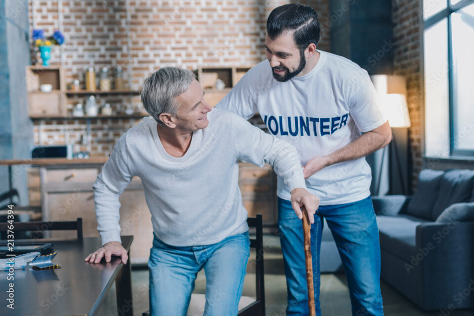 So helpful. Handsome joyful dark-eyed man smiling and helping an old man while he standing up